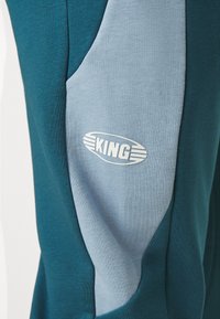 Pantaloni joggers di colore teal e azzurro chiaro con una texture morbida, caratterizzati da una vestibilità slim e un logo bianco "KING" in un design ovale sul lato inferiore.