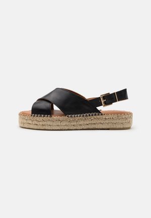 Sandalia espadrille de cuero negro con tiras cruzadas, hebilla en el tobillo y gruesa suela plataforma de yute tejido sobre fondo blanco.