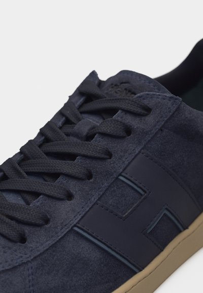 Gros plan sur une basket en daim marine avec des lacets noirs, un logo latéral et une semelle beige, sur un fond blanc uni.