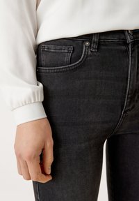 Jeans negros ajustados con textura desgastada y cinco bolsillos. Una camisa blanca de manga larga con puños elásticos es parcialmente visible arriba.