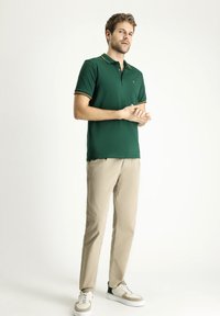 Polo verde con strisce contrastanti gialle e arancioni nel colletto, abbinato a pantaloni beige e sneakers bianche con accenti grigi e verdi.
