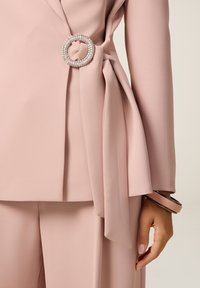 Lichtroze, op maat gemaakte outfit met een wikkeldesign en een met edelstenen versierde ronde broche die de taille accentueert, gecombineerd met een bijpassende armband.