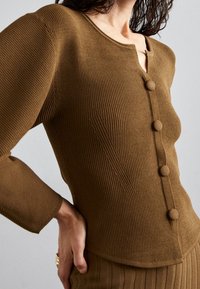 Brun ribbad stickad topp med rund halsringning, som har fem tygklädda knappar framtill och långa ärmar. Strukturerad yta.