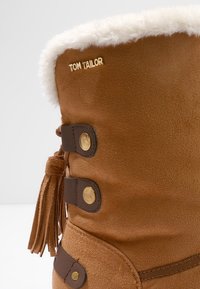 Brauner Wildleder-Winterstiefel mit weißem Kunstfellbesatz, drei Lederstephen mit Messingknöpfen, Quastendetail und "TOM TAILOR"-Logo.