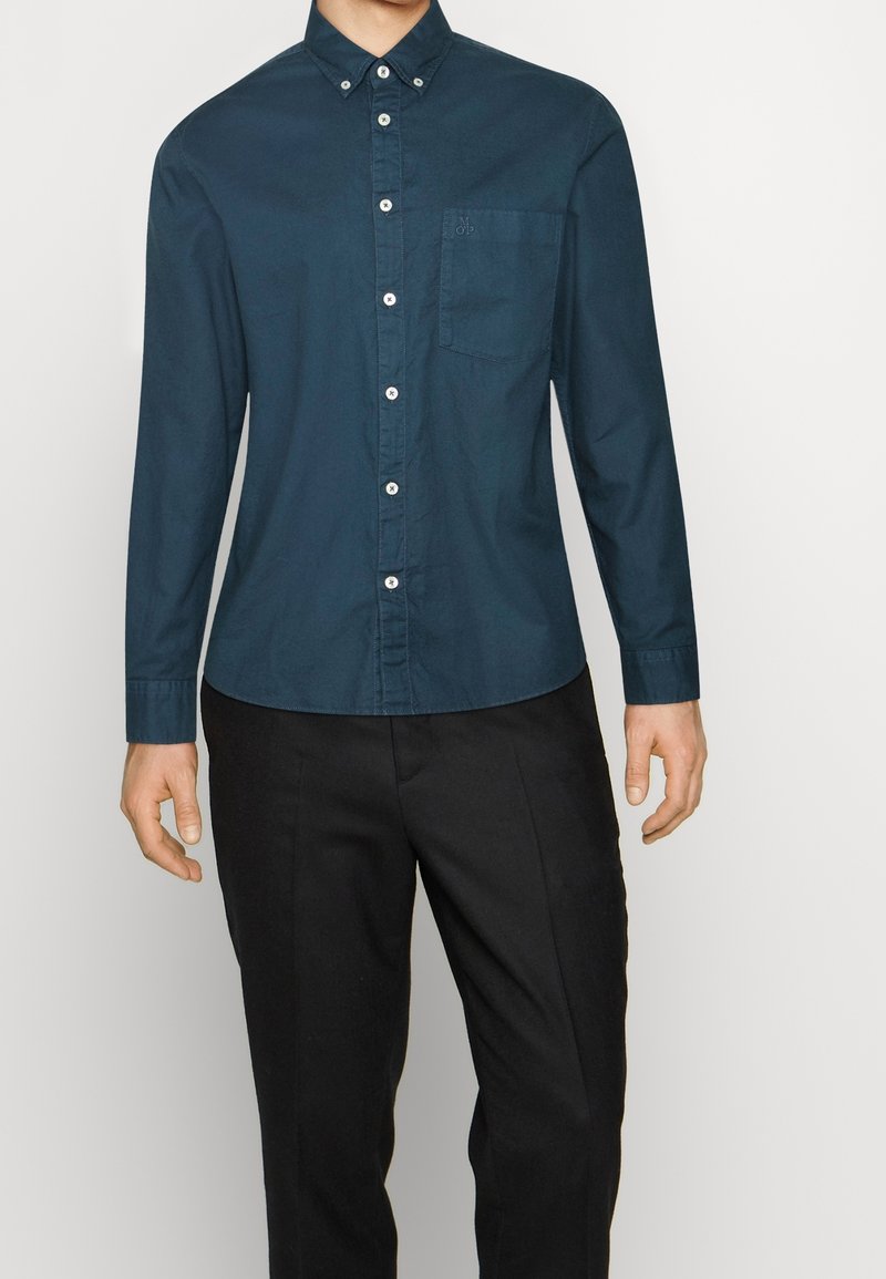 Homme portant une chemise bleu foncé boutonnée à manches longues avec une poche poitrine et un pantalon noir sur un fond clair uni.