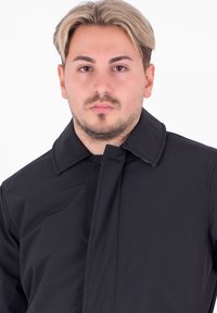 Cappotto nero con colletto alto, dotato di chiusura a un solo bottone, tessuto liscio e stile minimalista incentrato sulla funzionalità.