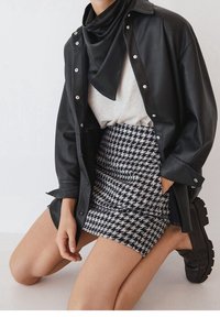 Personne à genoux portant une chemise en cuir noire, un bandana noir, un haut blanc, une jupe pied-de-poule noir et blanc, et des mocassins noirs épais.