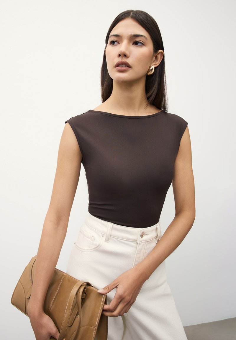 Woman wearing a sleeveless dark brown top and white pants, holding a tan leather handbag, looking slightly to the side against a white background.