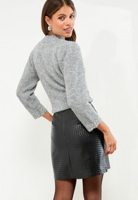 Szürke bolyhos pulóver háromnegyedes ujjakkal, gyöngy részletekkel a mandzsettán, egy fekete textúrált mini szoknyával párosítva.