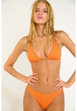 Femme aux longs cheveux blonds portant un bikini orange vif avec un pendentif en forme de cœur argenté, debout les mains sur les hanches devant un fond uni.