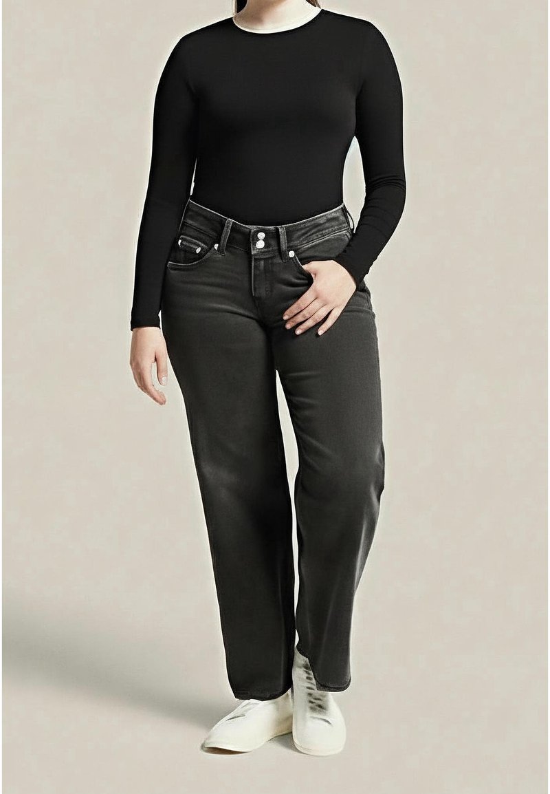 Personne portant un haut noir ajusté à manches longues, un jean gris foncé taille haute et des chaussures blanches, debout avec une main dans la poche.