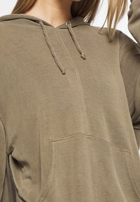 Olivgrön huvtröja med känguruficka. Tillverkad av mjukt material, har snodd i huven och ribbad muddar, med en avslappnad passform.