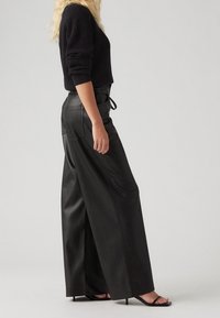 Czarne, szerokie skórzane spodnie z wysokim stanem, charakteryzujące się gładką strukturą, dwoma tylnymi kieszeniami oraz detalem w postaci sznurka w pasie.