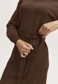 Vestito marrone lavorato a maglia con scollo a V, maniche lunghe e cintura annodata in vita. Presenta polsini a coste e una texture morbida.