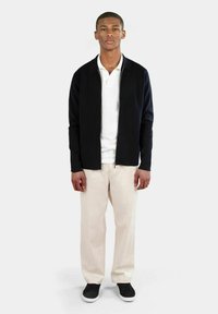 Svart zip-up jacka över en vit skjorta med krage, i kombination med ljusbeige byxor och svarta sneakers. Slätt tyg, avslappnad passform, inga mönster.