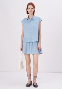 Light blue denim shirt with short sleeves, paired with a flared denim skirt. Model holds a small white handbag and wears black lace-up flats.