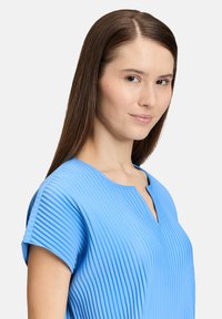 Jasnoniebieska plisowana bluzka z okrągłym dekoltem i krótkimi rękawami, z pionowymi teksturowanymi liniami oraz małym dekoltem w kształcie łezki z przodu.