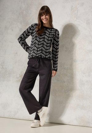 Black and white zigzag-patterned knit sweater, paired with black loose-fit trousers and white sneakers, against a textured gray background.