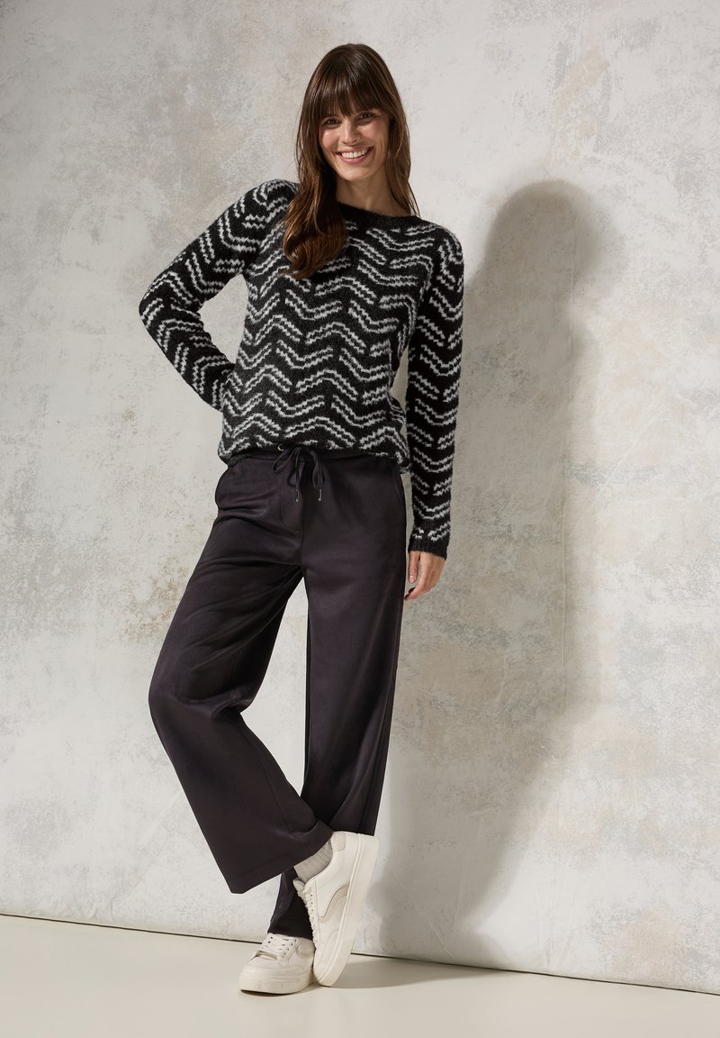 Pull en tricot noir et blanc à motif zigzag, associé à un pantalon noir décontracté et des baskets blanches, sur un fond gris texturé.