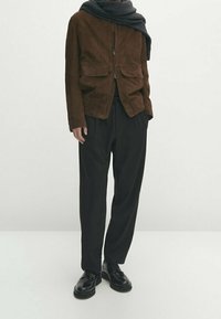 Bruine suède jas met twee voorzakken, gecombineerd met losse zwarte broek en zwarte veterschoenen. Een donkere gebreide sjaal wordt om de hals gedragen.