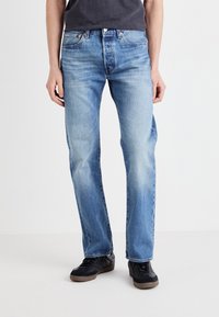 Jeans de mezclilla azul claro con corte recto y detalles de desvanecimiento. Llevados con zapatillas negras que tienen una suela texturizada.