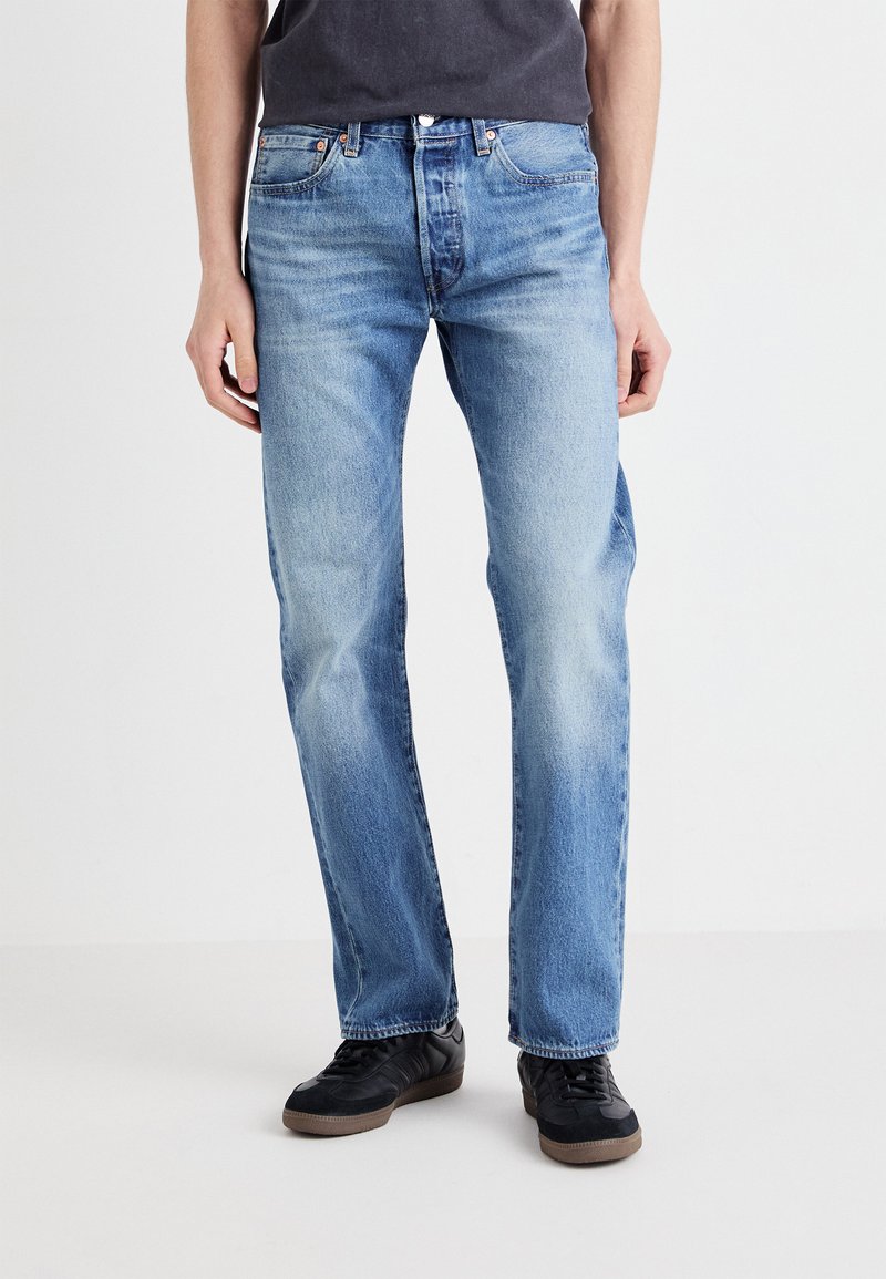 Jeans de mezclilla azul claro con corte recto y detalles de desvanecimiento. Llevados con zapatillas negras que tienen una suela texturizada.