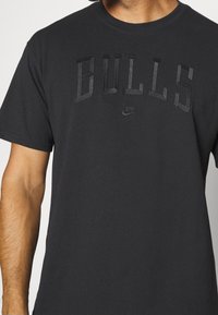 Černé bavlněné tričko s nápisem "BULLS" v texturovaných tmavých písmenech a malým logem Nike níže. Kulatý výstřih a krátké rukávy.