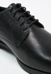 Chaussure oxford noire en cuir avec une texture lisse, un bout rond et un système de laçage. Elle présente des coutures discrètes et un petit talon.