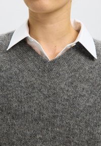 Person som har på seg en hvit skjorte med krage under en grå, fluffy strikket genser med V-hals, som viser nakken og den nedre delen av ansiktet.