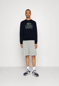 Mornarska sweatshirt z zelenim Lacoste logotipom, v kombinaciji s svetlo sivo kratkimi hlačami. Model nosi bele nogavice in mornarske superge s kremnimi akcenti.