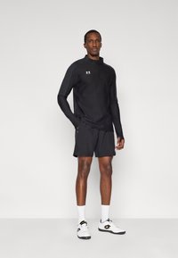 Μαύρο αθλητικό μπλουζάκι με φερμουάρ και μακριά μανίκια, μαύρο σορτς και λευκά αθλητικά παπούτσια. Διαθέτει το λογότυπο της Under Armour και υφασμάτινες λεπτομέρειες στους ώμους.