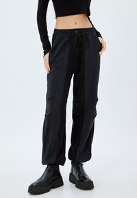 Schwarze, locker sitzende Hose mit strukturiertem Oberflächenmaterial, ausgestattet mit einem elastischen Bund und Kordelzug, kombiniert mit robusten schwarzen Stiefeln.