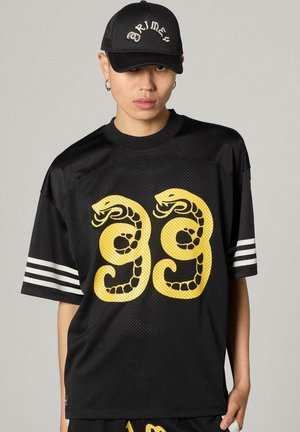 Młoda osoba ubrana w czarny sportowy meshowy strój z dwoma żółtymi zwiniętymi węzami na przodzie oraz czarną czapkę z napisem "Grimes".