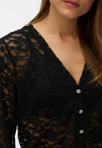 Blusa in pizzo nero con scollo a V, caratterizzata da motivi floreali, tessuto trasparente e decorata con bottoni argentati sul davanti.