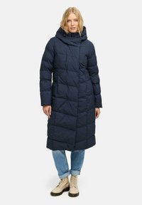 Marinblå lång pufferjacka med hög krage och huva. Har ett quiltat m�önster, dragkedjestängning och vida ärmar. Kombinerad med ljusblå jeans och beige stövlar.