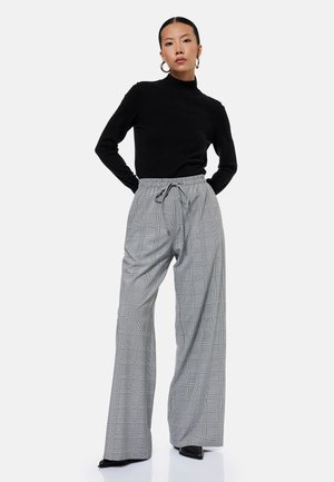 Kvinde iført sort rullekrave, store hoop-øreringe og højtaljede grå ternede vidde bukser står med hænderne bag ryggen.