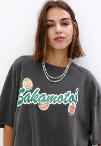 Šedé oversized tričko s nápisem "Sakamoto's" v zelené barvě, zdobené vícebarevnými kulatými nálepkami. Vícebarevný korálkový náhrdelník.