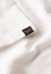 Witte stof met een zachte textuur en een lichte glans. Bevat een zwart label met "SUPERDRY" in oranje letters, genaaid aan de rand.