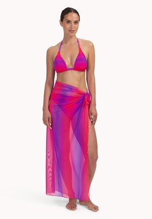 Femme portant un bikini dégradé rose et violet avec un paréo transparent assorti noué à la taille, debout pieds nus sur un fond blanc.
