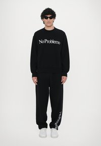 Svart hettegenser med teksten "No Problemo", kombinert med matchende svarte joggebukser med den samme teksten. Hvite sneakers fullfører antrekket.