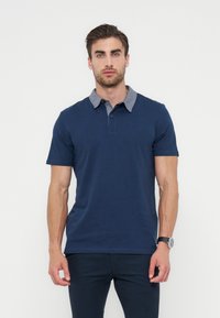 Άνδρας που φοράει μπλε σκούρο πόλο με γκρι γιακά και σκούρο παντελόνι, στέκεται μπροστά σε απλό λευκό φόντο.