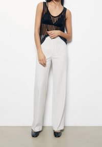 Haut débardeur noir transparent à rayures verticales associé à un pantalon blanc taille haute à jambes larges. Mannequin portant des chaussures noires brillantes.