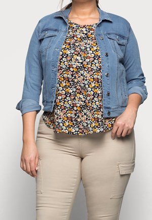 Kvinde iført opkørt denimjakke, blomstret top med gule og hvide blomster og beige bukser med sidelommer, står mod en ensfarvet baggrund.