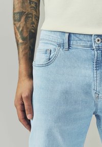 Jeans in denim azzurro chiaro con vestibilità slim, dotati di tasche frontali, cuciture a contrasto e chiusura con bottone metallico. Texture liscia.