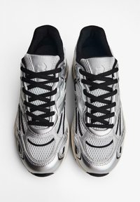 Des chaussures de sport argentées présentent une tige en mesh et synthétique avec des lacets noirs, des motifs texturés et une semelle en caoutchouc pour l'adhérence.
