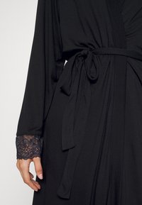 Robe noire à manches longues, avec une bordure en dentelle aux poignets et un détail de taille nouée. Le tissu fluide drape élégamment.