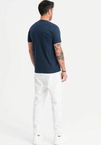 Marinefarvet bomulds T-shirt med korte ærmer og rund hals, parret med hvide bukser og hvide sneakers. Synlige tatoveringer på armen.