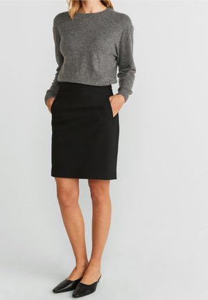 Mujer vestida con un suéter gris de manga larga, falda negra hasta la rodilla con bolsillos y zapatos negros sin cordones con punta puntiaguda, de pie.