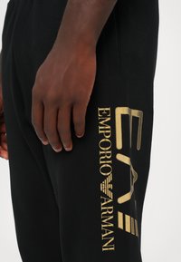 Fekete sportnadrág, amelynek oldalán egy feltűnő arany "Emporio Armani" logó található. Az anyag simának tűnik, és testhezálló sziluettet formál.