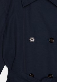 Trench-coat bleu marine en tissu lisse, avec un design à double boutonnage, de grands boutons noirs et une taille ceinturée pour un ajustement réglable.